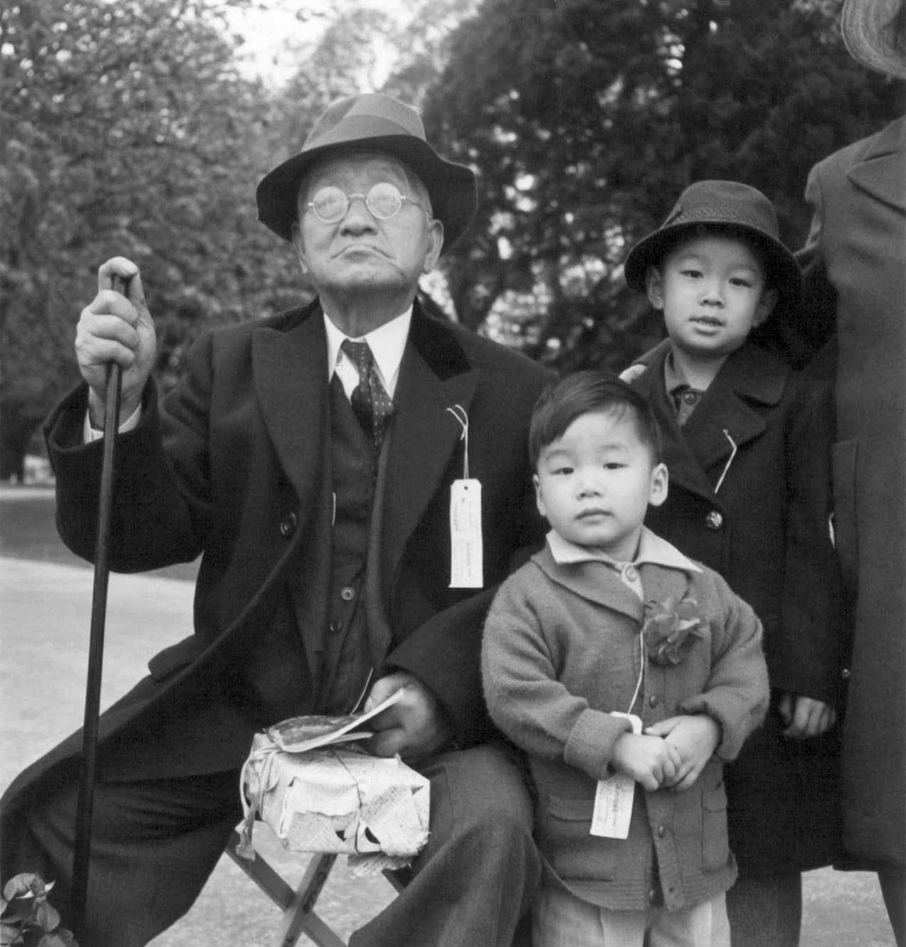 A grandfather and his grandchildren await an evacuation bus in 1942, in Hayward, California, during the Japanese evacuation and internment (Dorothea Lange Photo).