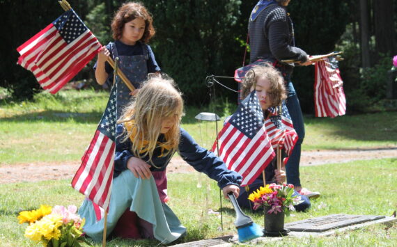 Tom Hughes Photo
Members of Vashon’s Cub Scout Pack 275, Luna Cekosh (kneeling, left), Hazel Ecevedo (standing), and Wilfred Gogarten (right) place American flags on the graves of veterans at Vashon Cemetery.