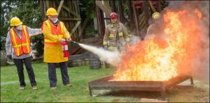 Practical Training with Fire Extinguishers: New CERT volunteers Joni Larned (left) and Kate OHare (with extinguisher) buddy up to get experience using a fire extinguisher. Vashon Island Fire & Rescue (VIFR) Captain Josh Munger (left rear) and volunteer firefighter Jim Wescott (right rear) instructed the volunteers at the VIFR training ground.