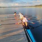 Rose Ely, Gwen Tomlinson, Ren Colvos, Emily Rock, and Alekos Dalinis sit with rows at the ready. Coach Delany Steele crouches on the pier nearby (Ben Steele photo).