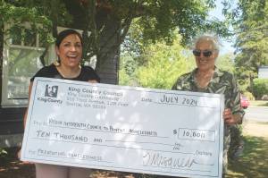 King County Councilmember Teresa Mosqueda (left) holds a big novelty check with the Vashon Interfaith Council to End Homelessness' Hilary Emmer, outside the Chamber of Commerce, on July 31. (Alex Bruell photo)