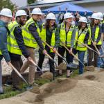 Local and regional leaders ceremonially strike shovels into the dirt at a ground-breaking ceremony for the Vashon-Maury Food Banks new location at the United Methodist Church, as a crowd behind raises apples celebratorily. (Alex Bruell photo)